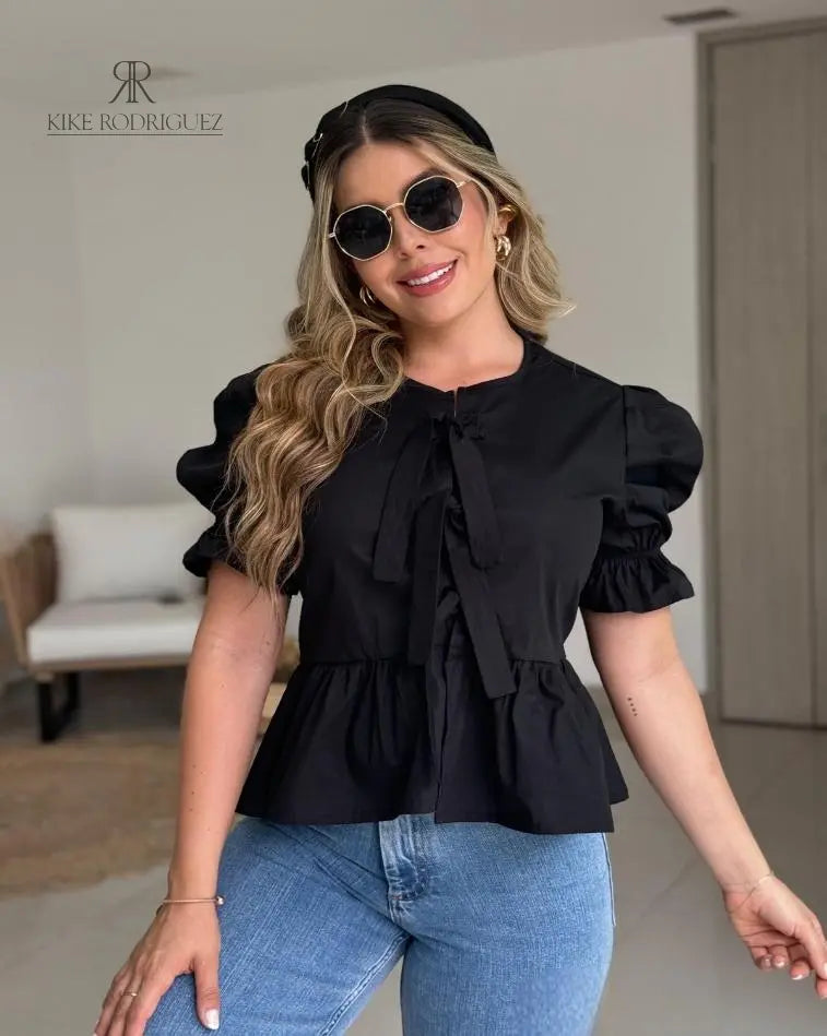 Mujer vistiendo la Blusa Matilda negra con diseño peplum, mangas abullonadas y lazos frontales, combinada con jeans para un look casual y moderno
