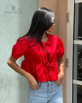 Mujer vistiendo la Blusa Matilda Roja con diseño peplum, mangas abullonadas y lazos frontales, combinada con jeans para un look casual y moderno