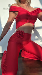 Mujer vistiendo un conjunto rojo sexy de KIKE RODRIGUEZ, con un top off-shoulder y pantalones acampanados, mostrando un look elegante y atractivo bajo la luz del sol.