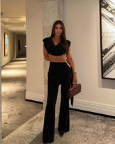 Mujer usando el Set Atenea Negro, con top corto y pantalón de tiro alto, posando en pasillo elegante con cartera de mano vinotinto.