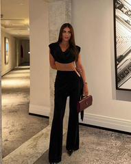 Mujer usando el Set Atenea Negro, con top corto y pantalón de tiro alto, posando en pasillo elegante con cartera de mano vinotinto.