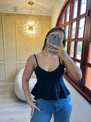 Mujer vistiendo un top peplum negro de tirantes, posando frente a un espejo con un entorno elegante de fondo.