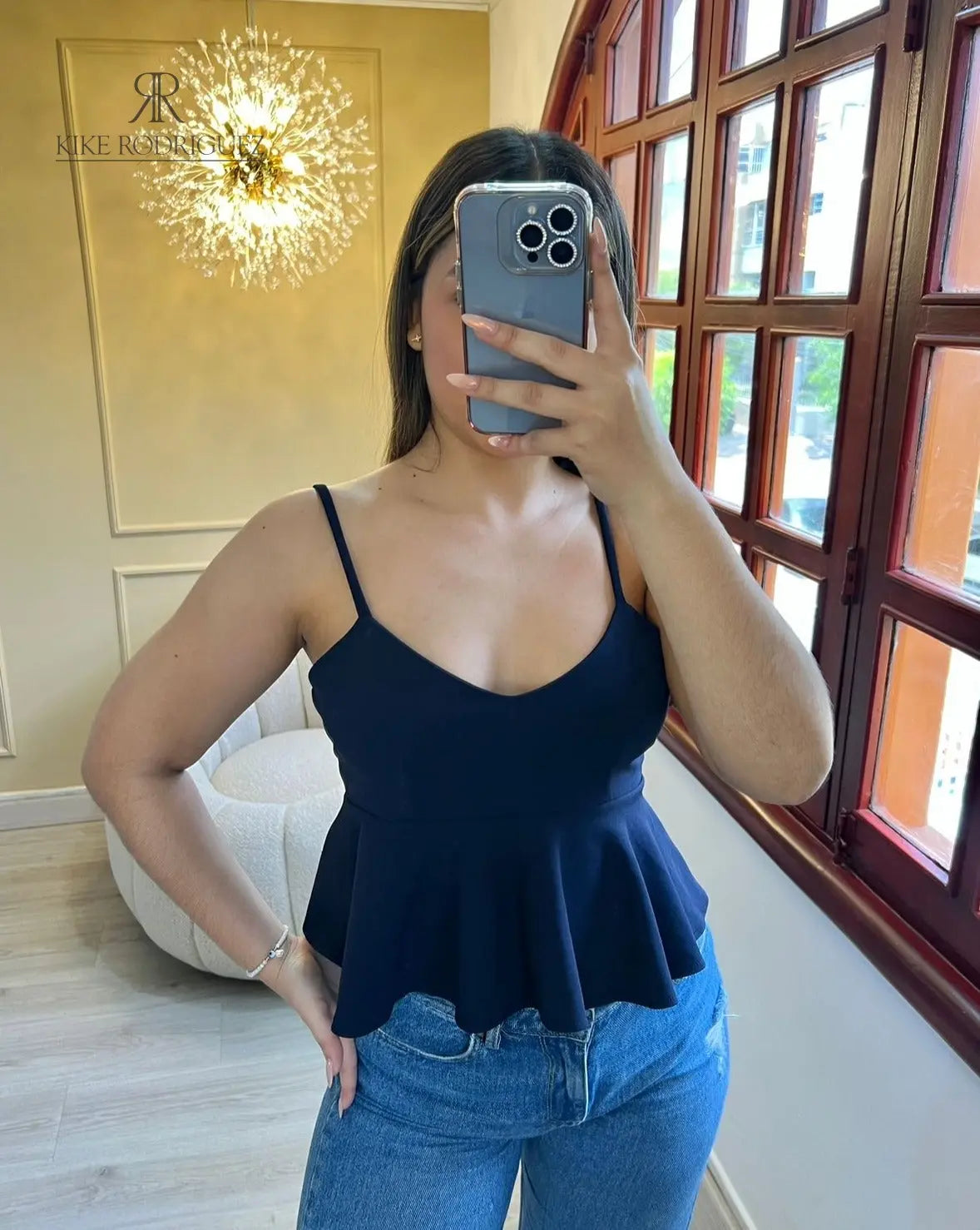 Mujer vistiendo un top peplum negro de tirantes, posando frente a un espejo con un entorno elegante de fondo.