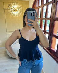 Mujer vistiendo un top peplum negro de tirantes, posando frente a un espejo con un entorno elegante de fondo.