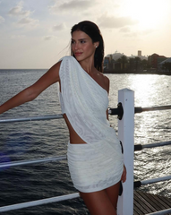 Mujer en muelle al atardecer usando el Set Taurus Little Skirt blanco de un solo hombro, con fondo de mar y luz dorada.