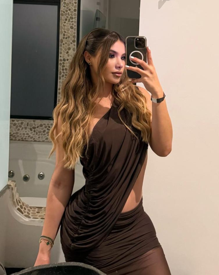 Mujer posando frente al espejo con el Set Taurus MOCCA, vestido marrón con corte lateral y escote asimétrico, peinado con ondas y maquillaje natural.