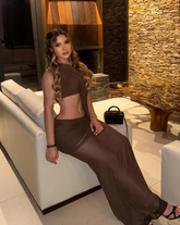 Mujer luciendo el Set Taurus MOCCA en tono marrón, de espalda, en un ambiente moderno con paredes de piedra y muebles blancos. Vestido ajustado con diseño asimétrico y apertura lateral.