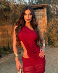 Mujer luciendo el Set Taurus VINOTINTO en tono vinotinto, de espalda, en un ambiente moderno con paredes de piedra y muebles blancos. Vestido ajustado con diseño asimétrico y apertura lateral.