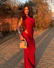 Mujer luciendo el Set Taurus VINOTINTO en tono vinotinto, de espalda, en un ambiente moderno con paredes de piedra y muebles blancos. Vestido ajustado con diseño asimétrico y apertura lateral.