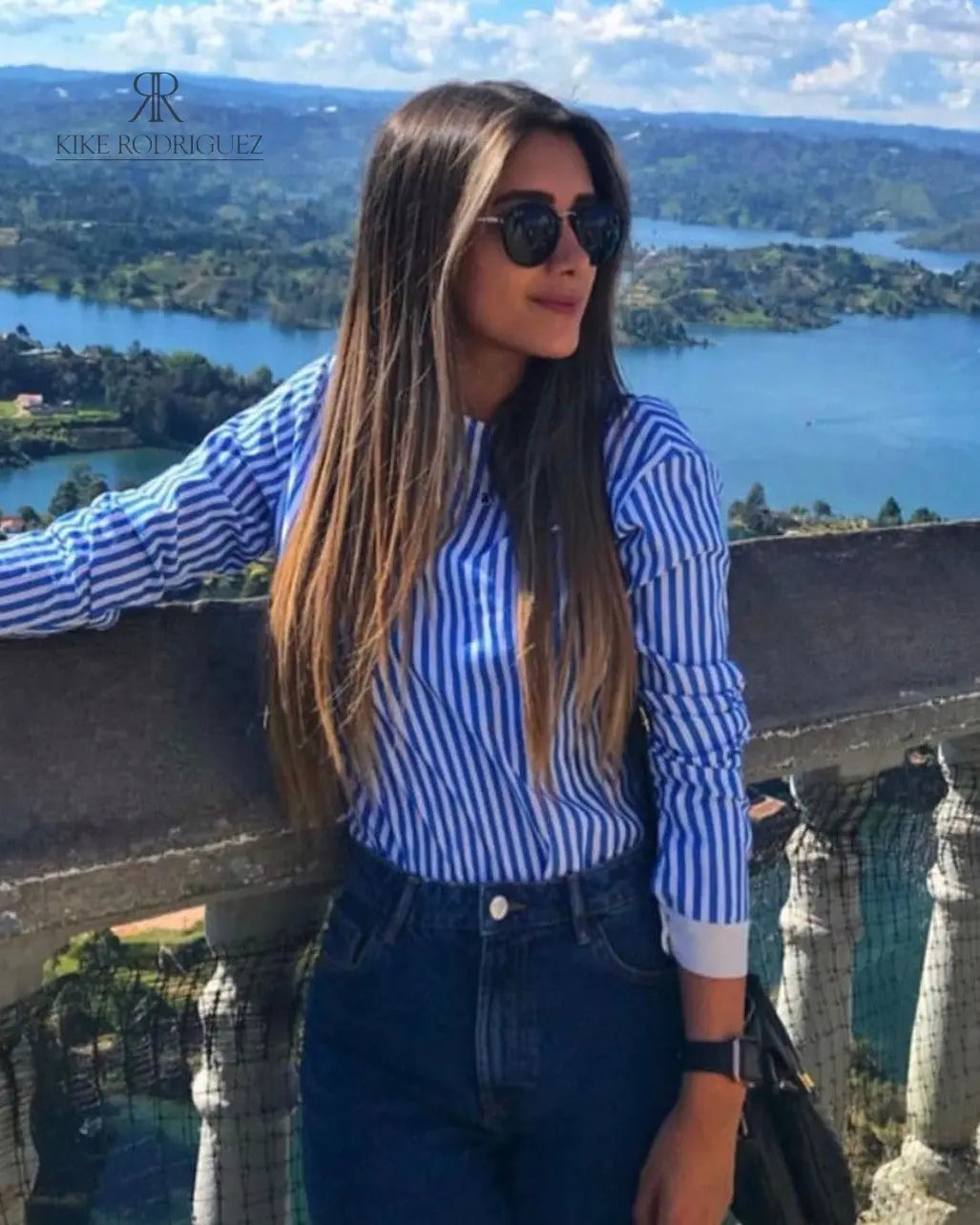Mujer con blusa de rayas con espalda descubierta disfrutando de una vista panorámica. Blusa perfecta para un look casual y sofisticado. Disponible en KIKE RODRIGUEZ.