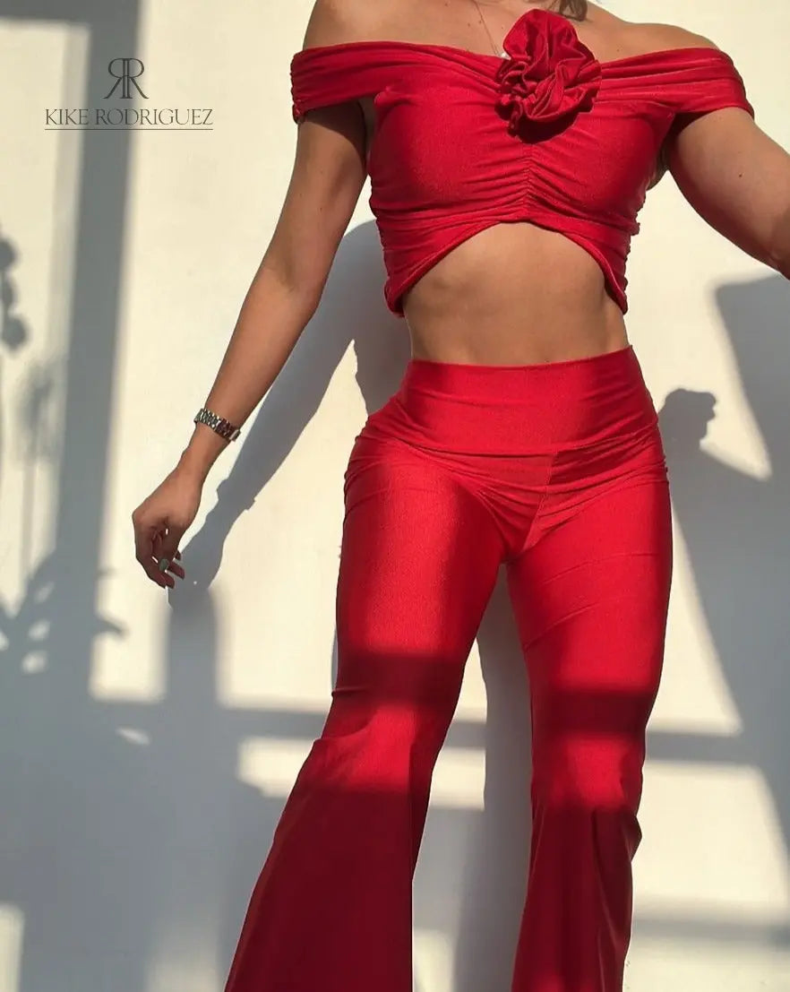 Mujer vistiendo un conjunto rojo sexy de KIKE RODRIGUEZ, con un top off-shoulder y pantalones acampanados, mostrando un look elegante y atractivo bajo la luz del sol.