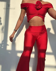 Mujer vistiendo un conjunto rojo sexy de KIKE RODRIGUEZ, con un top off-shoulder y pantalones acampanados, mostrando un look elegante y atractivo bajo la luz del sol.