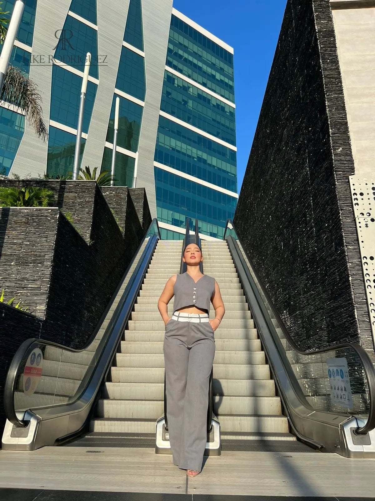 Mujer vistiendo un conjunto de pantalón y chaleco gris de KIKE RODRIGUEZ, posando en una escalera moderna con un fondo arquitectónico y vegetación.