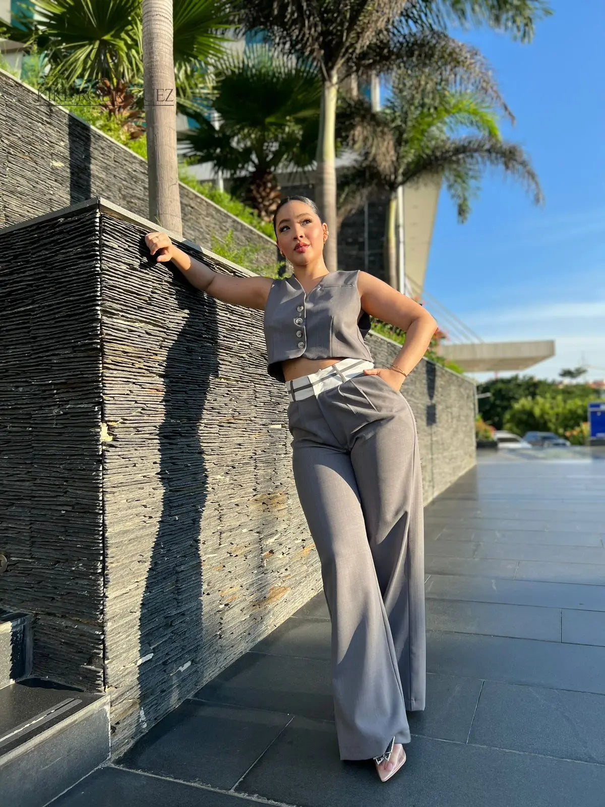 Mujer vistiendo un conjunto de pantalón y chaleco gris de KIKE RODRIGUEZ, posando en una escalera moderna con un fondo arquitectónico y vegetación.