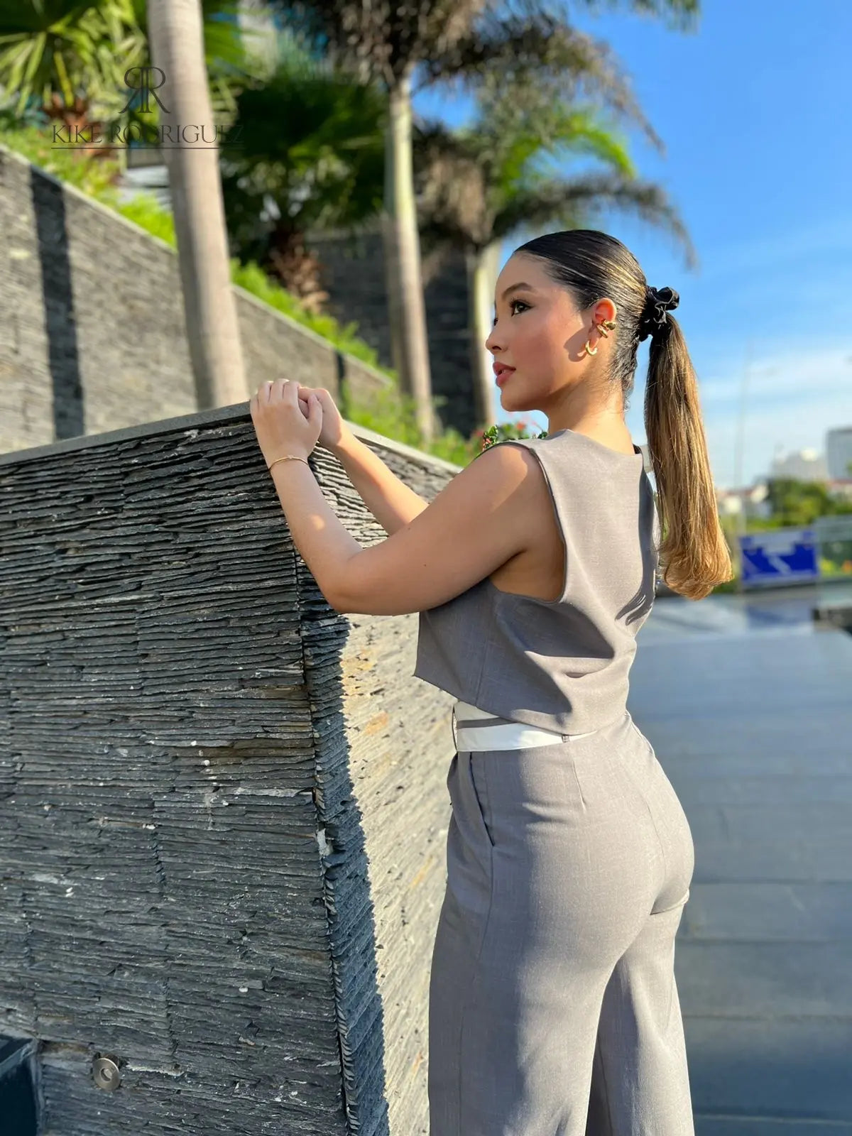 Mujer vistiendo un conjunto de pantalón y chaleco gris de KIKE RODRIGUEZ, posando en una escalera moderna con un fondo arquitectónico y vegetación.