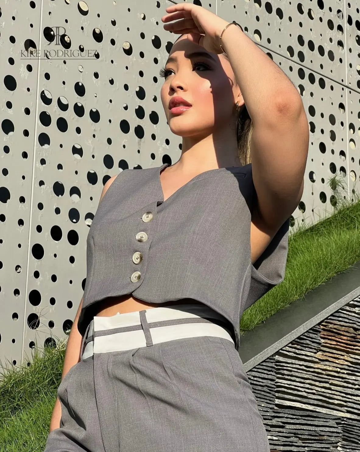 Mujer vistiendo un conjunto de pantalón y chaleco gris de KIKE RODRIGUEZ, posando en una escalera moderna con un fondo arquitectónico y vegetación.