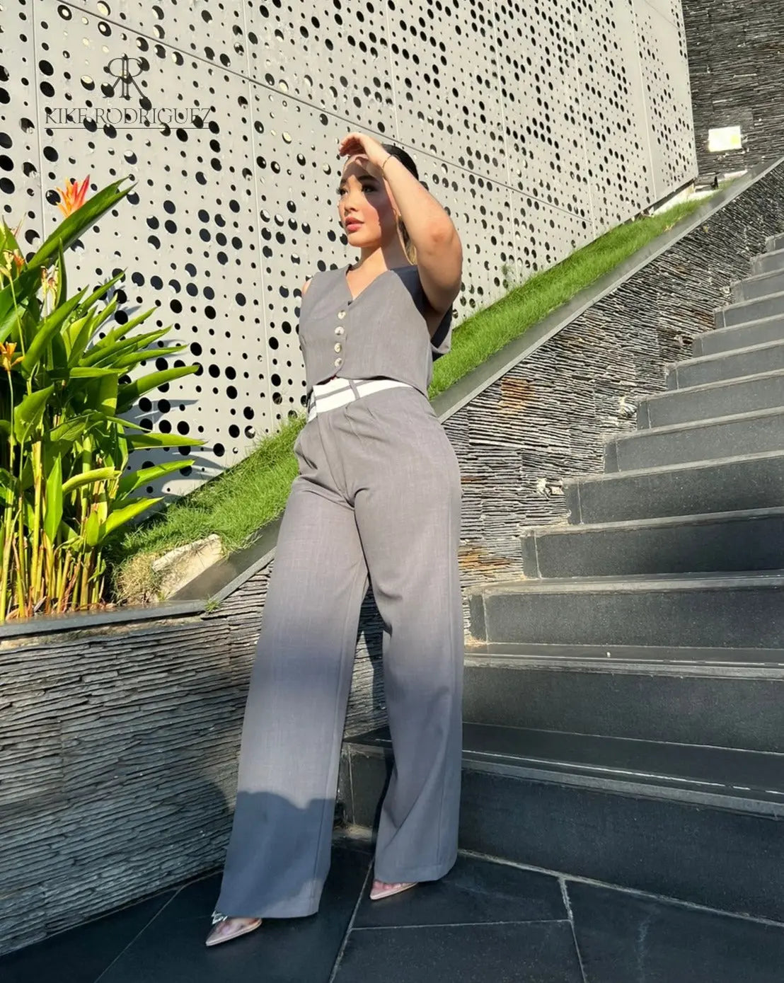 Mujer vistiendo un conjunto de pantalón y chaleco gris de KIKE RODRIGUEZ, posando en una escalera moderna con un fondo arquitectónico y vegetación.