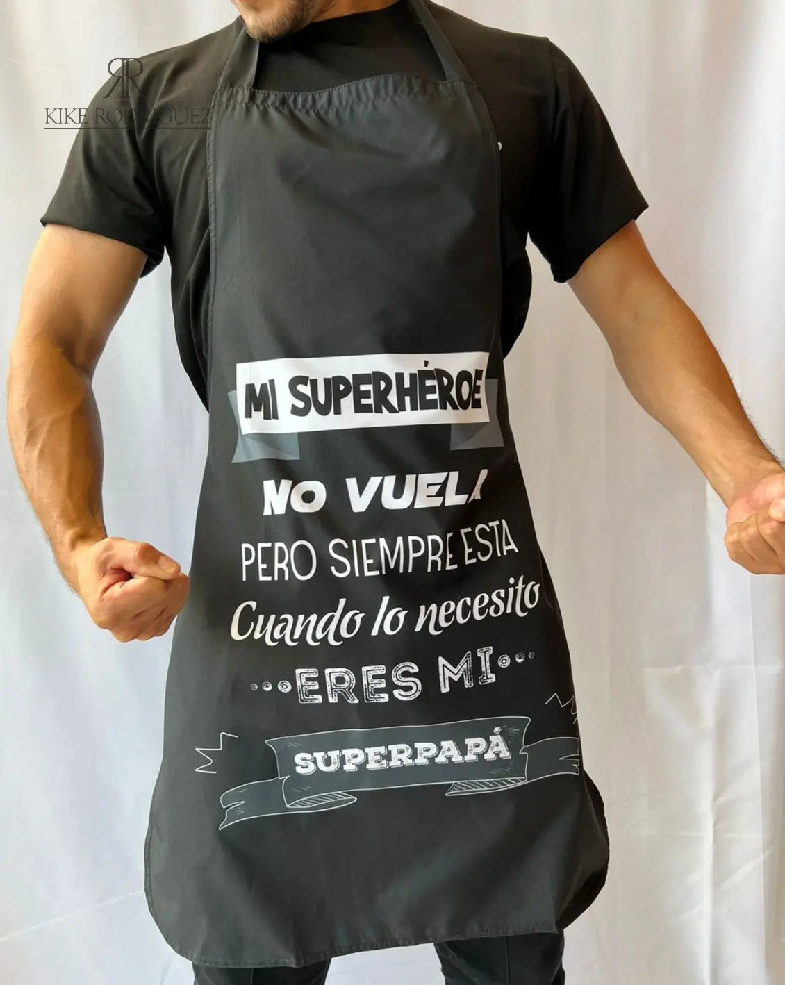 Hombre usando un delantal de cocina personalizado en color negro con el mensaje "Mi superhéroe no vuela, pero siempre está cuando lo necesito. Eres mi superpapá".