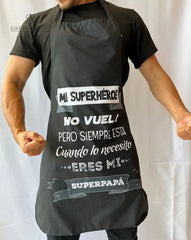 Hombre usando un delantal de cocina personalizado en color negro con el mensaje "Mi superhéroe no vuela, pero siempre está cuando lo necesito. Eres mi superpapá".