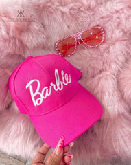 Gorra rosa de Kike Rodriguez con el logo de Barbie en blanco, acompañada de gafas de sol rosas sobre un fondo de peluche, ideal para un look femenino, moderno y divertido