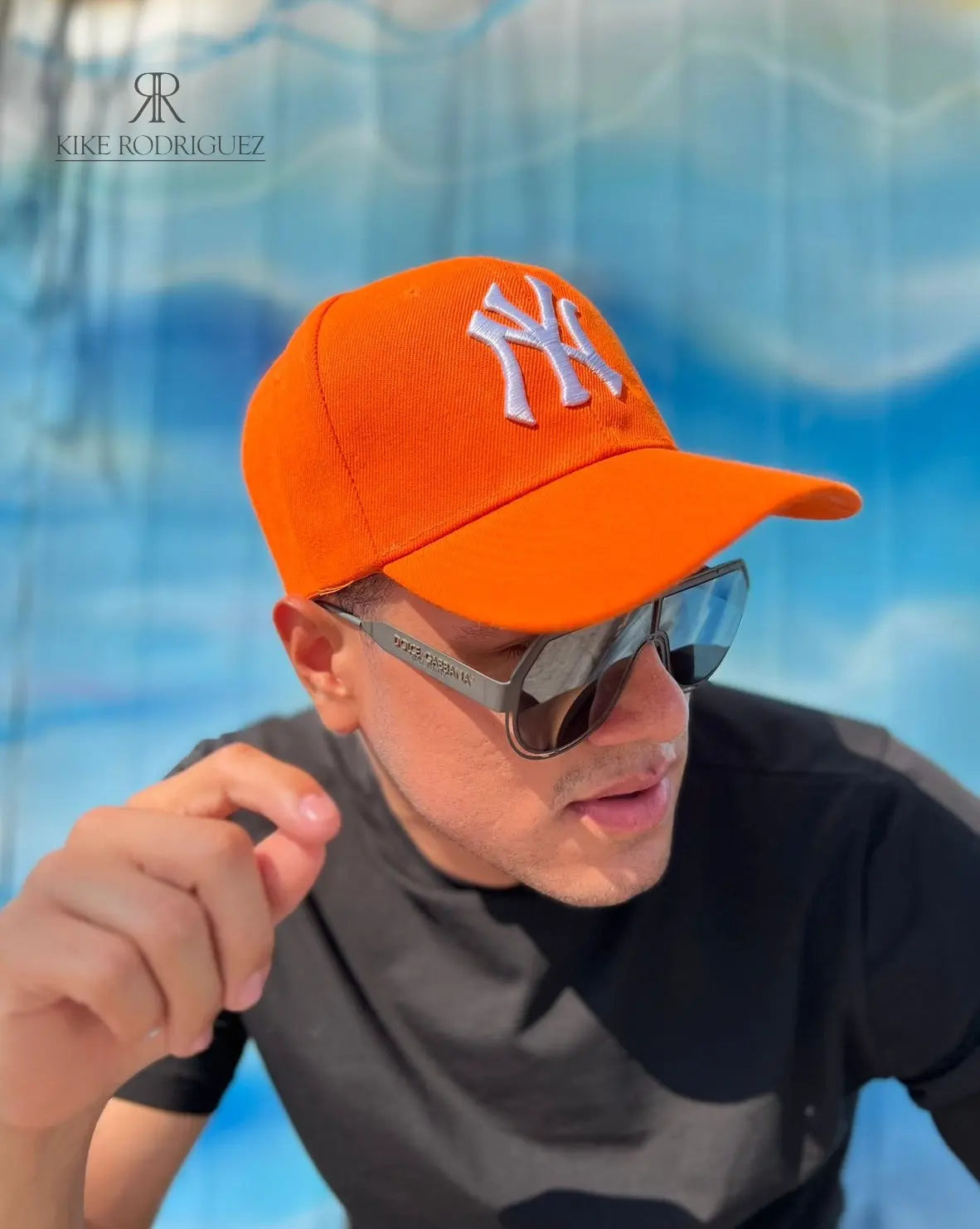 Hombre usando una gorra naranja de Kike Rodriguez con logo blanco de los New York Yankees, combinada con gafas de sol y camiseta negra para un look deportivo y moderno