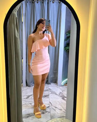 Mujer vistiendo un elegante vestido asimétrico rosa claro con volantes, posando frente a un espejo en un entorno moderno y sofisticado.