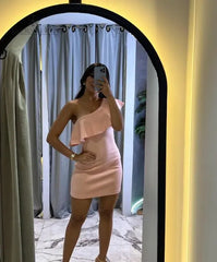 Mujer vistiendo un elegante vestido asimétrico rosa claro con volantes, posando frente a un espejo en un entorno moderno y sofisticado.