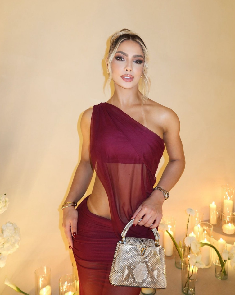 Mujer luciendo el Set Taurus VINOTINTO en tono vinotinto, de espalda, en un ambiente moderno con paredes de piedra y muebles blancos. Vestido ajustado con diseño asimétrico y apertura lateral.